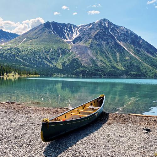 A canoe is on the shore of a clear teal lake with a large mountain in the background