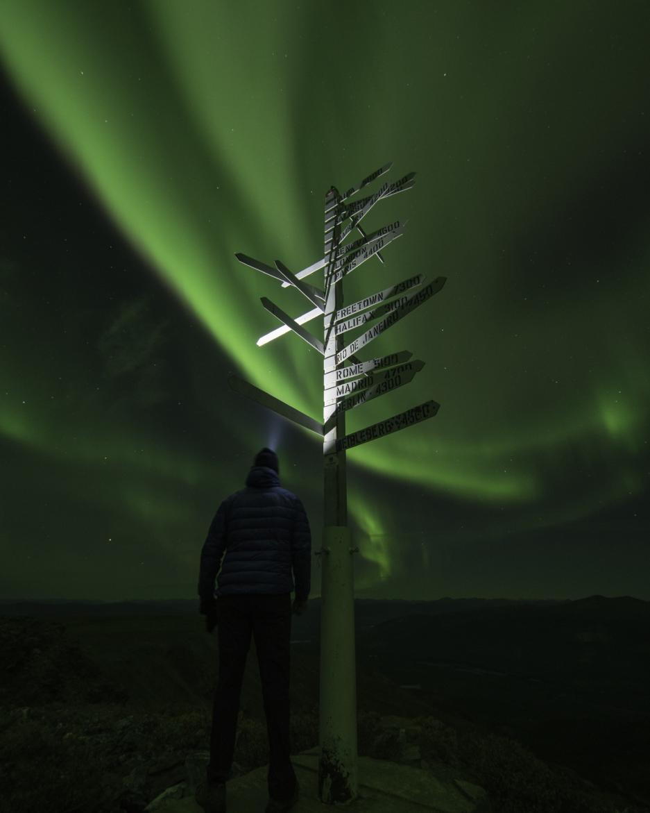 A person stands beside a white sign post under the aurora